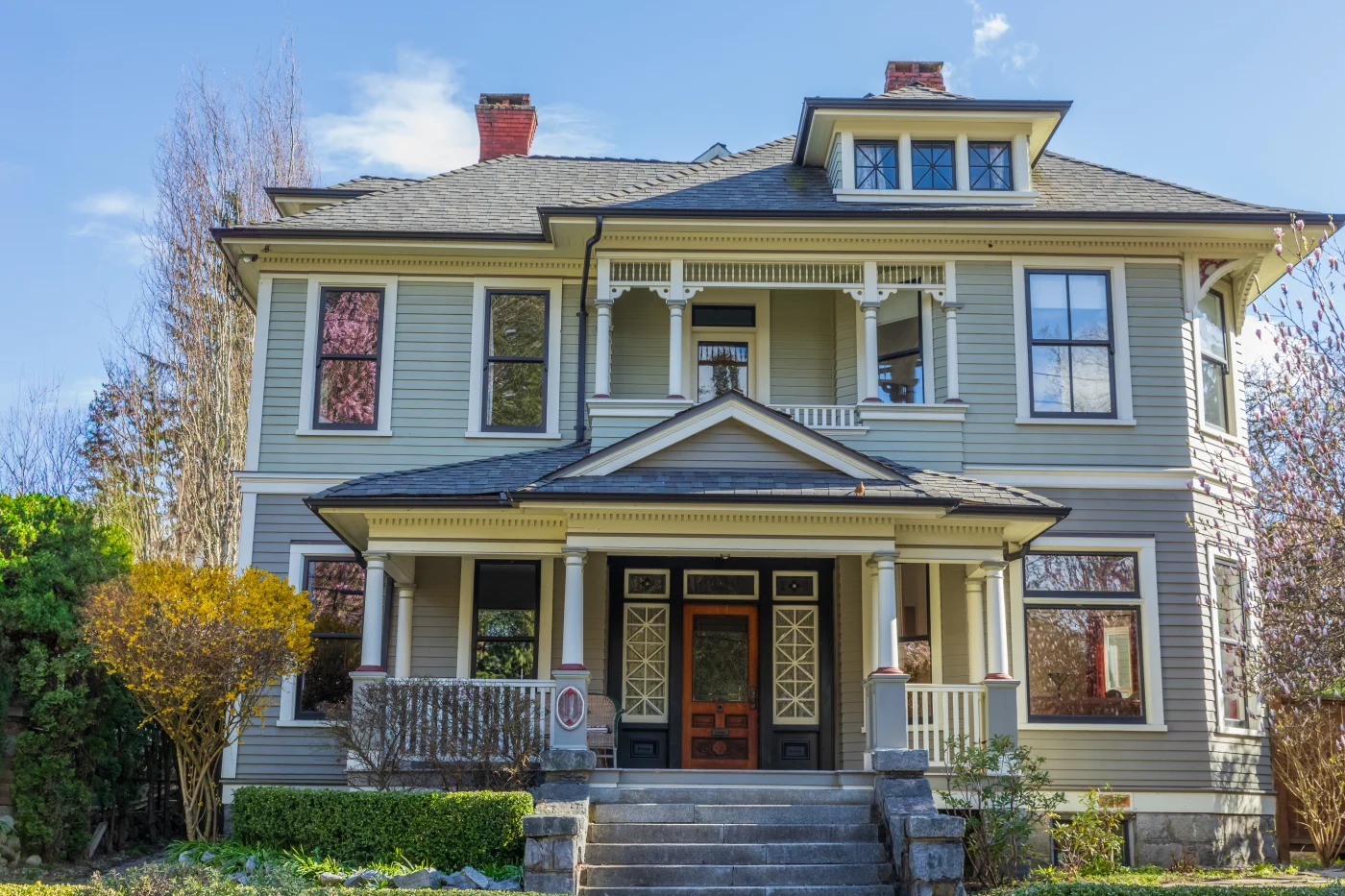 An upward view of a tan three-story house with cement steps, a wooden front door, blue windows, a second-story balcony, and an asphalt shingle roof.