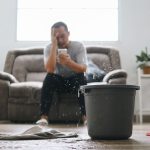 A picture of a man sitting on a couch, holding his phone in one hand and covering half of his face with the other. In the foreground, a gray bucket and white towel are visible on the floor, and water is dripping down from the ceiling.
