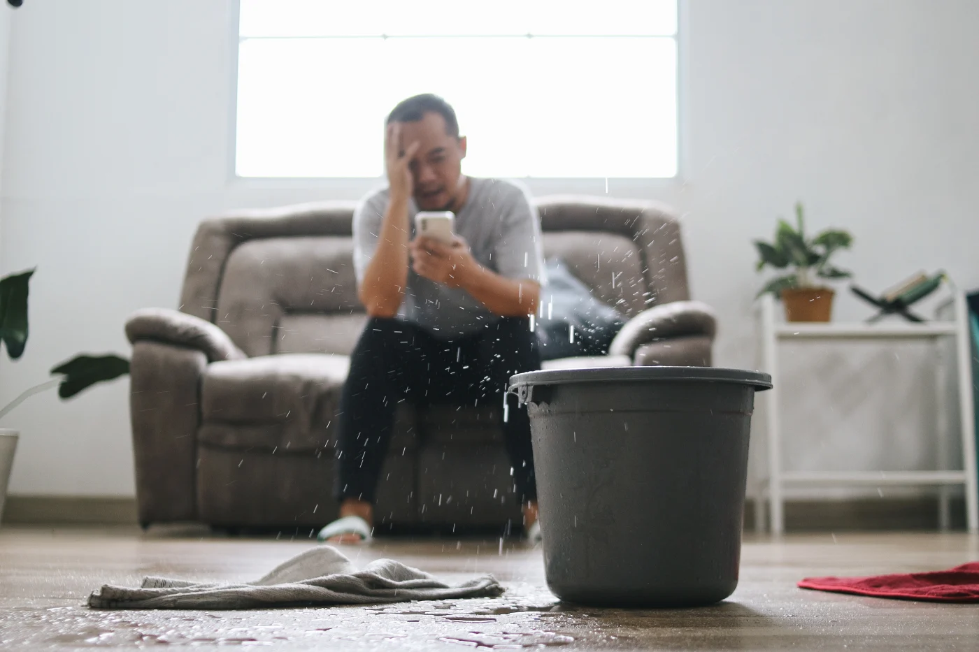 Welcome Roof Repair Menasha Wi Wisconsin Exteriors A picture of a man sitting on a couch, holding his phone in one hand and covering half of his face with the other. In the foreground, a gray bucket and white towel are visible on the floor, and water is dripping down from the ceiling.