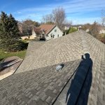 A picture taken on top of a roof, from one end. The photographer’s shadow is visible on the surface, as are the shingles, which are various shades of gray. Beyond the home, another house is visible, as is the yard, and a few tall green trees.