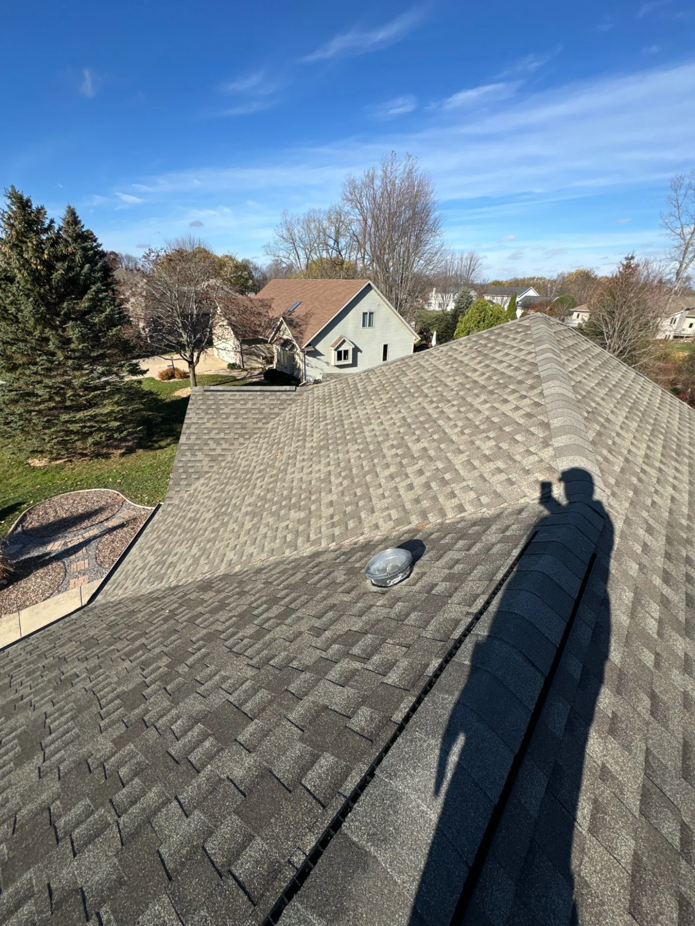 Wholesome Roof Inspection Menasha Wi Wisconsin Exteriors A picture taken on top of a roof, from one end. The photographer’s shadow is visible on the surface, as are the shingles, which are various shades of gray. Beyond the home, another house is visible, as is the yard, and a few tall green trees.